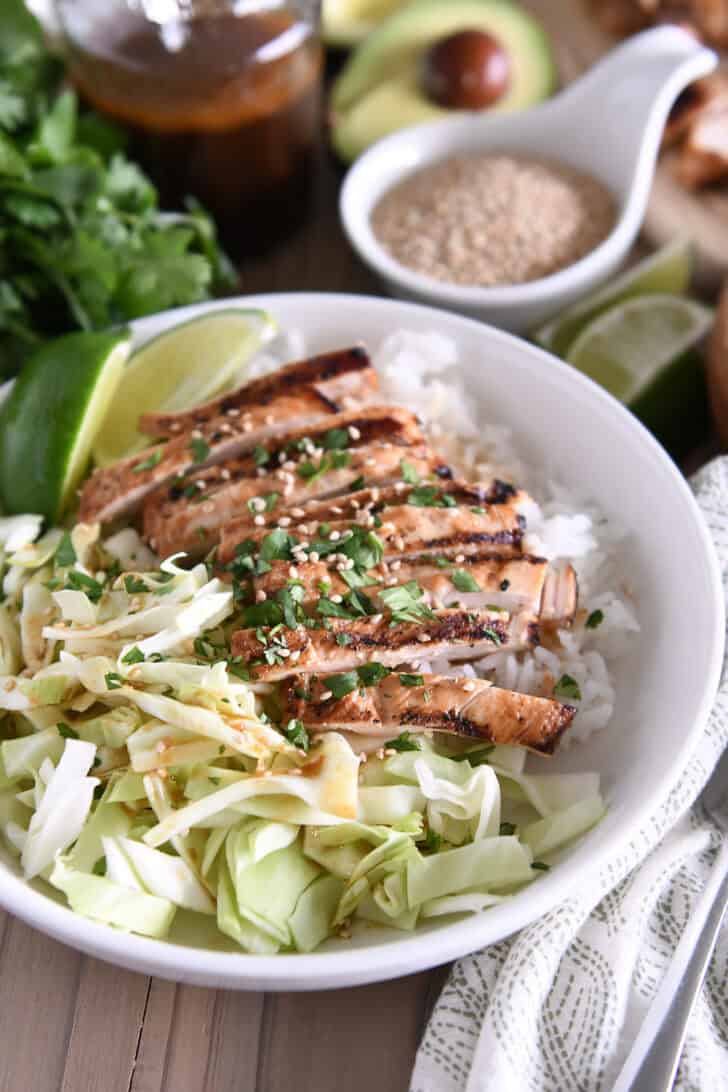 White bowl with chopped cabbage, white rice, grilled chicken, lime wedges, chopped cilantro and toasted sesame seeds.