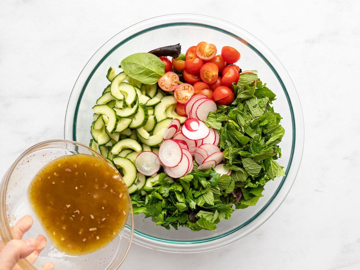 pouring dressing over greens, mint, parsley, tomatoes, cucumber, bell pepper and radishes in large bowl