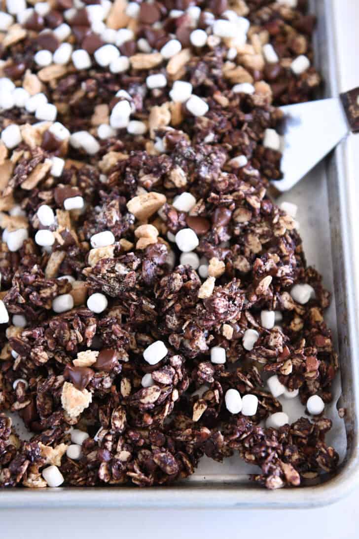 Chocolate granola on parchment-lined sheet pan with graham cracker pieces and mini marshmallows and chocolate chips.