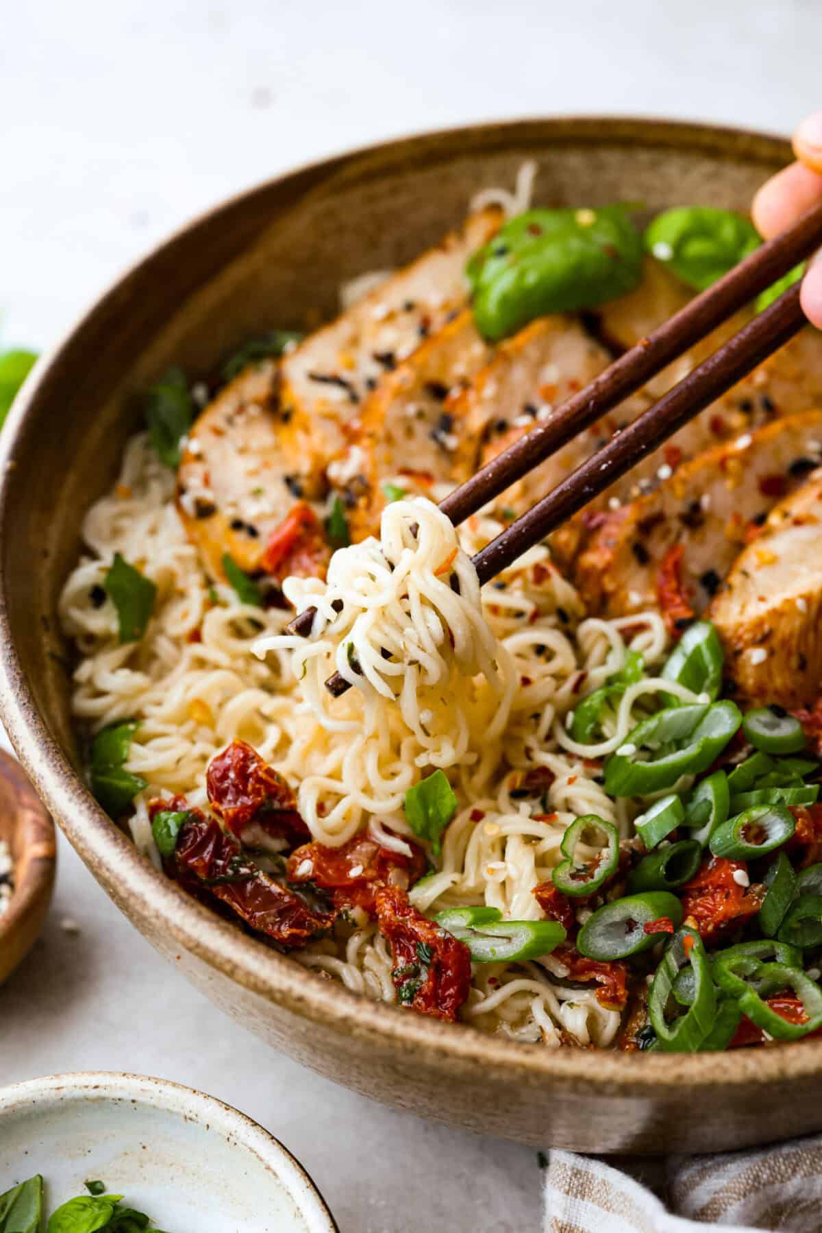A bowl of creamy Marry Me Ramen with tender noodles, grilled chicken, sun-dried tomatoes, and fresh green onions. Chopsticks lifting noodles from the rich, flavorful broth.