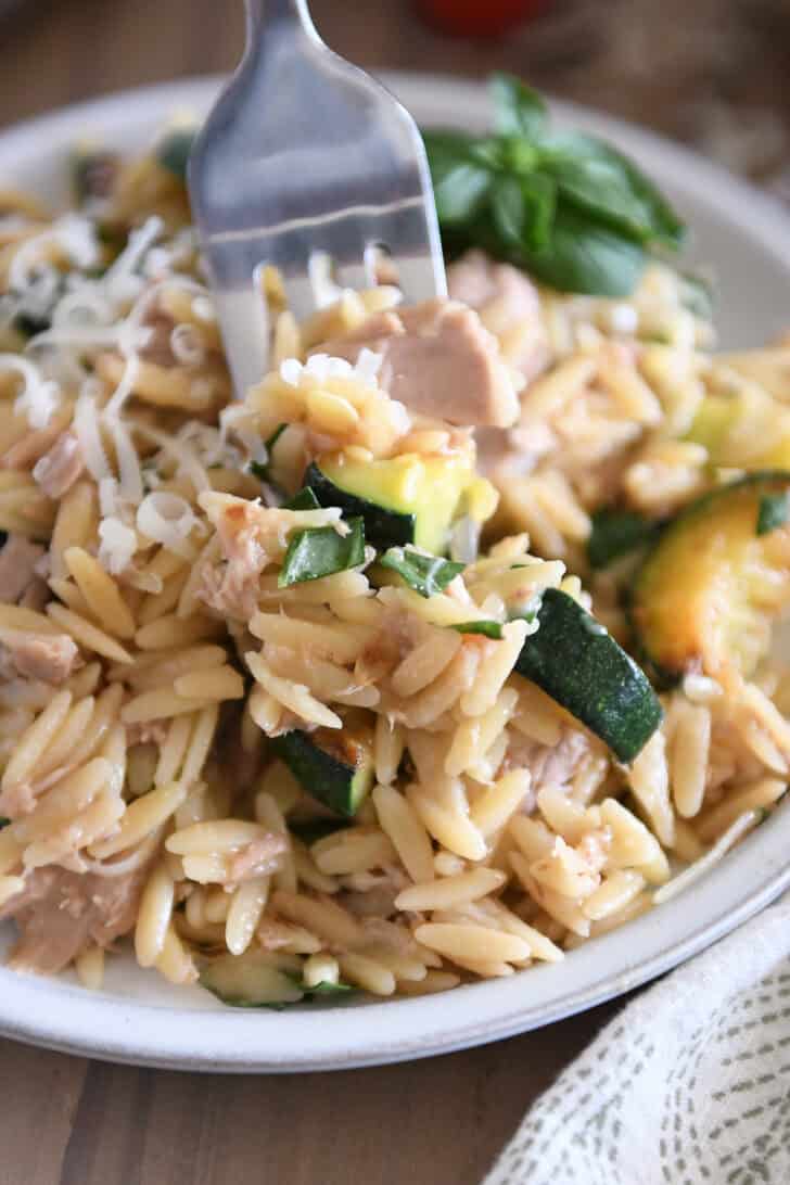 Orzo with tuna, zucchini, and Parmesan with fresh basil as a garnish on gray plate and fork scooping bite.