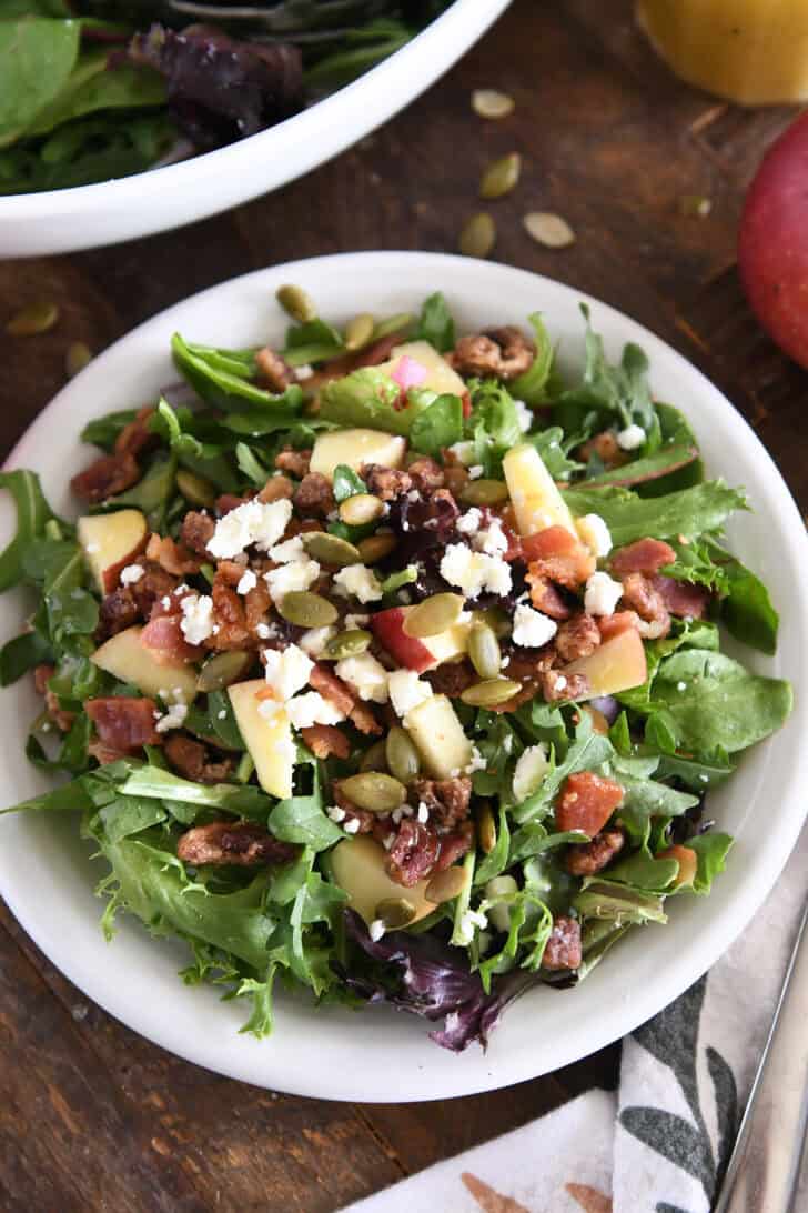 Top down view of green salad on white plate with roasted pumpkin seeds, chopped apples, candied pecans, feta cheese and red onions.