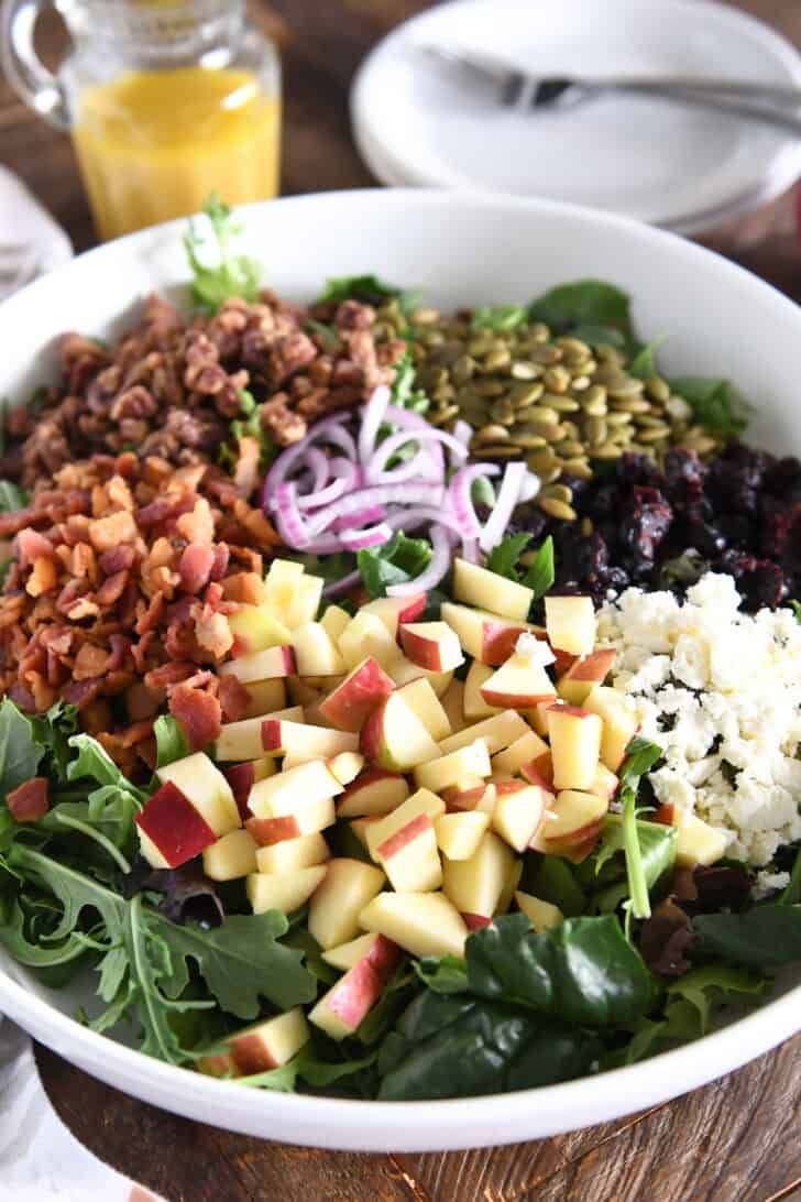 White bowl with chopped apples in the foreground and feta cheese, bacon, candied pecans, roasted pumpkin seeds and red onions in the background.