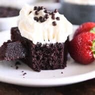 Piece of chocolate olive oil cake topped with whipped cream and crispy chocolate pearls with bite of cake on fork on white plate.