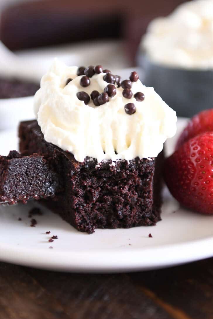 Piece of chocolate olive oil cake topped with whipped cream and crispy chocolate pearls with bite of cake on fork on white plate.