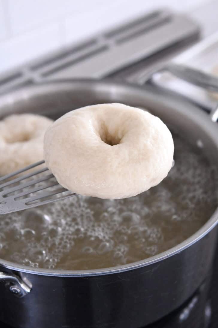 Lifting boiled bagel out of the water with stainless spatula.