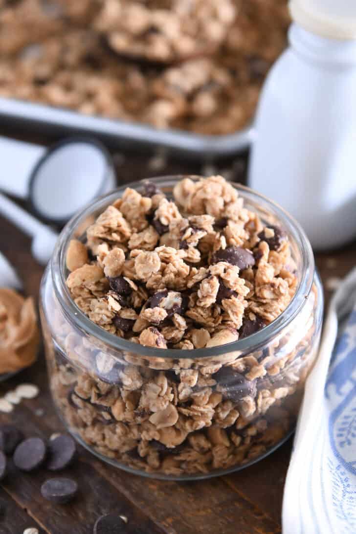 Shallow glass jar filled with peanut butter granola with roasted peanuts and dark chocolate chunks with spoonful of peanut butter to the side.