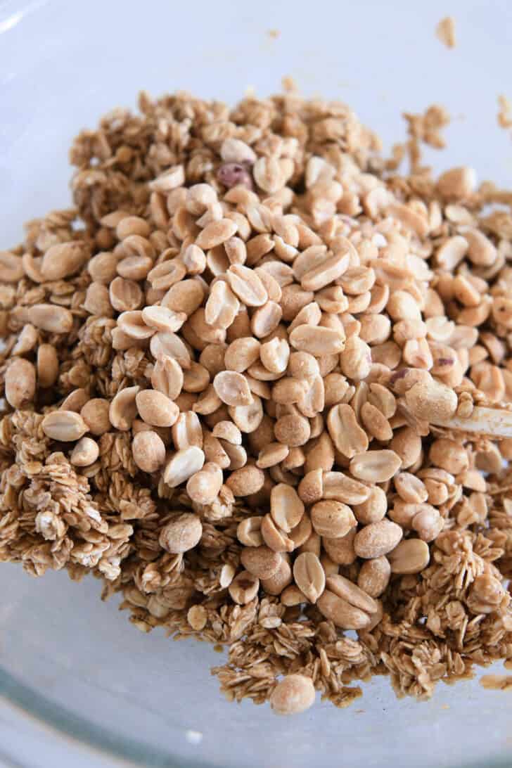 Roasted peanuts added on top of wet peanut butter granola mixture in glass bowl.
