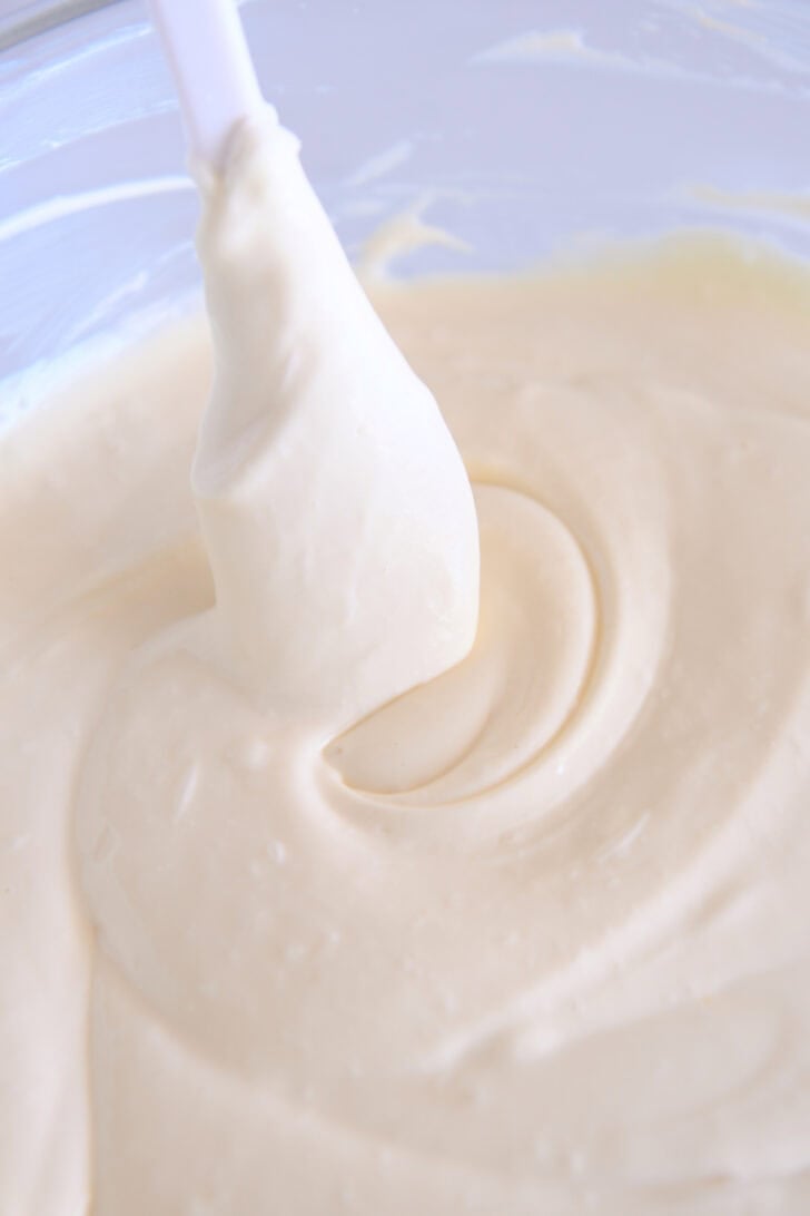 White spatula stirring cheesecake batter in glass bowl.