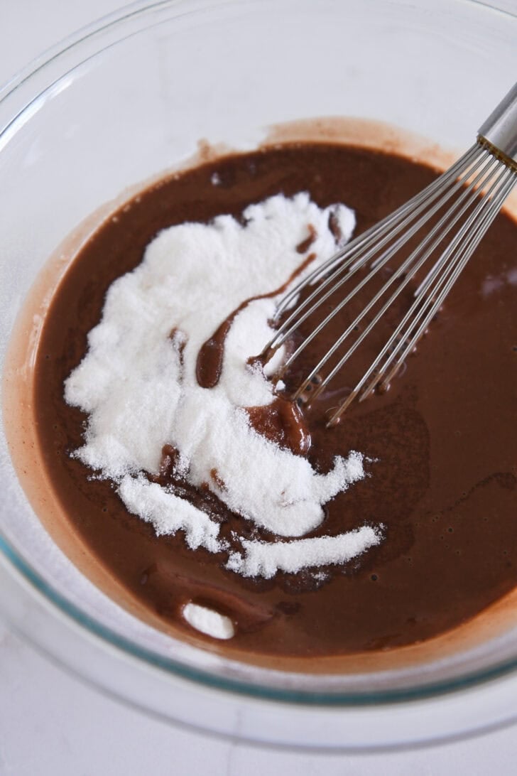 Glass bowl with liquidy chocolate mixture, granulated sugar, and wire whisk.