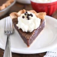 Triangle slice of chocolate chess pie on white plate with fork to the side and a dollop of whipped cream on top of pie topped with chocolate crisp pearls.