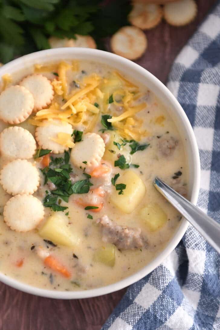 Soup bowl with sausage potato soup topped with oyster saltine crackers, fresh parsley and grated cheese and spoon on side.