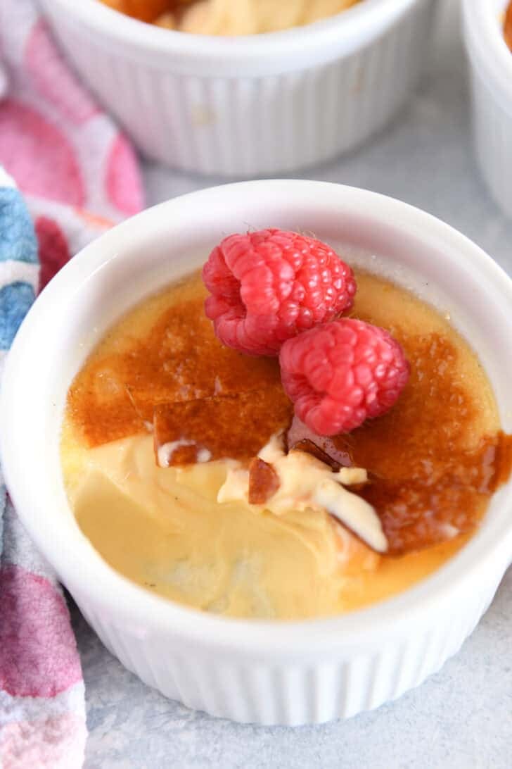 Scoop taken out of creme brûlée in white ramekin with raspberries on the side.