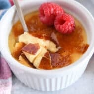 Spoon cracking through top shell of creme brûlée in white ramekin with raspberries on the side.