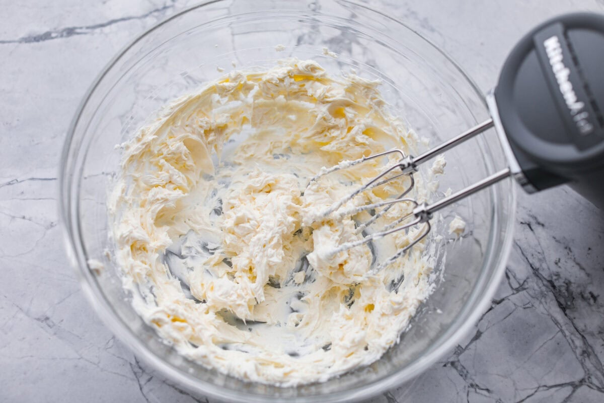 Someone using a hand mixer to beat the cream cheese in a mixing bowl.