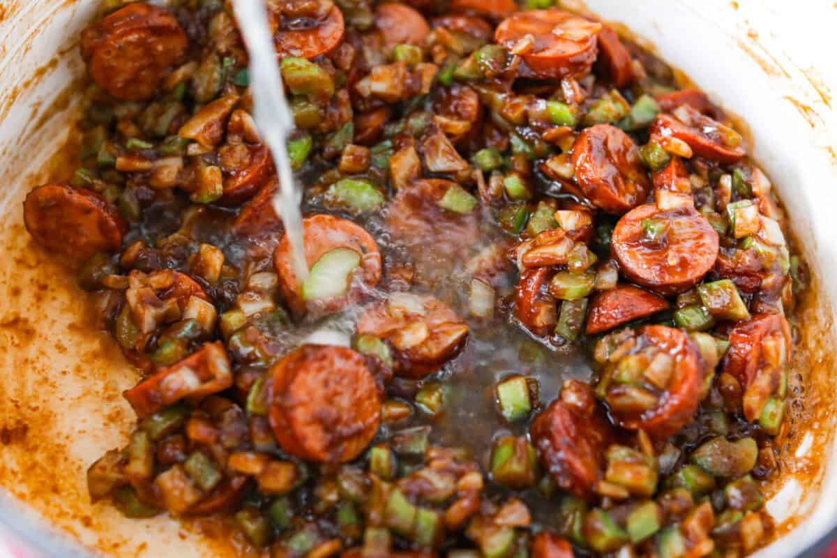 Someone slowing pouring chicken broth into the pot with the sausage veggies and roux.