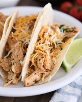 A white plate with three shredded chicken tacos topped with cheese in flour tortillas and a lime wedge next to the tacos.