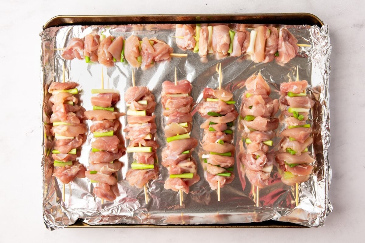foil-lined sheet pan with uncooked chicken and scallion pieces threaded onto skewers