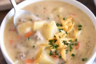 White bowl with spoon scooping ingredients in creamy cheesy soup with cooked potatoes, ground beef, and carrots and topped with shredded cheddar cheese and chopped chives.