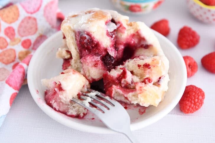 Fork taking piece of raspberry sweet roll on white plate.