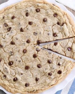 Giant chocolate chip cookie on white platter with slice cut.