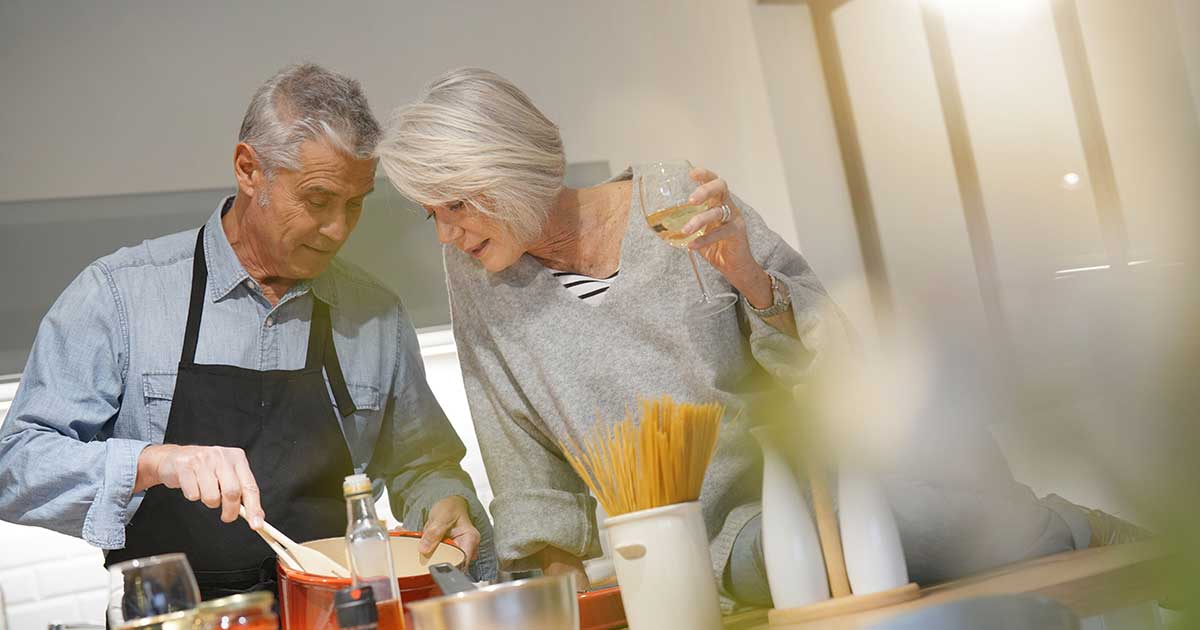 Couple cooking together — both actively participating, him chopping, her stirring, wine glasses, teamwork visible