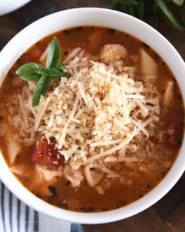 Top down view of chicken parmesan soup with cooked pasta, chicken, tomatoes, and seasonings in white bowl topped with buttered bread crumbs with Parmesan.