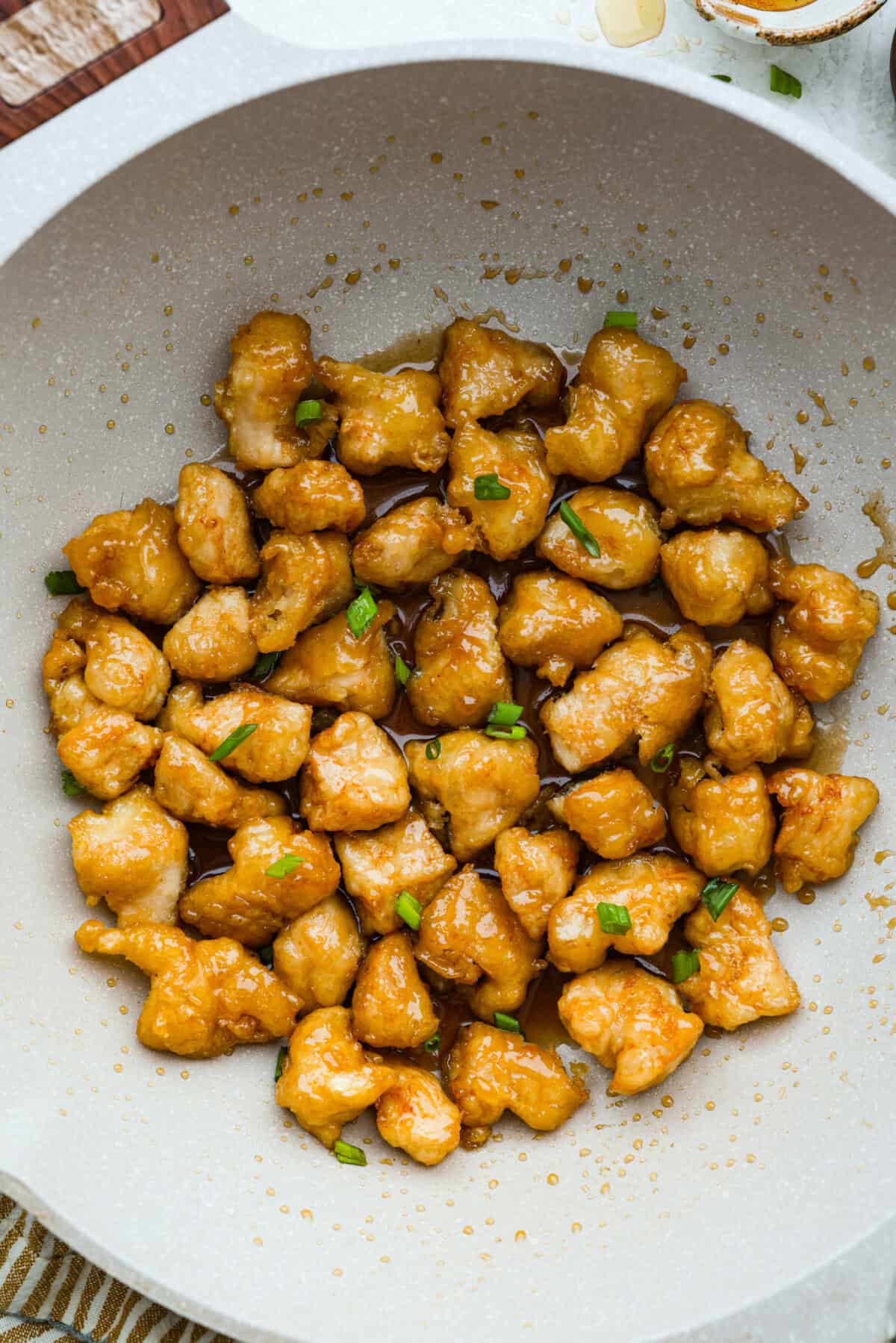 Top down shot of a wok with fried chicken pieces covered in the honey sauce and garnished with green onions. 