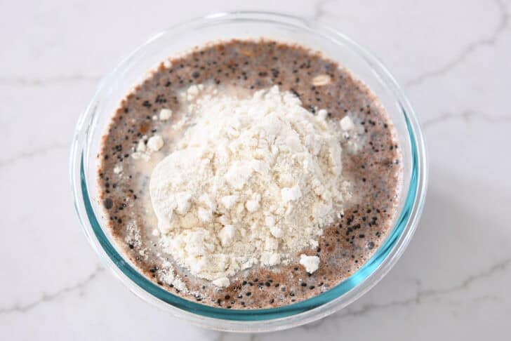 Glass bowl with almond milk, chia seeds, basil seeds, oats, cinnamon, maple syrup, protein powder, and salt.