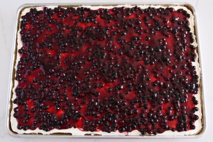 White sheet cake in half sheet pan topped with cream cheese frosting and huckleberry fruit topping.