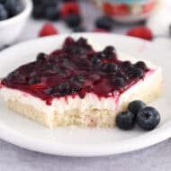 Square of white sheet cake topped with cream cheese layer and fruit topping on white plate with fresh blueberries.