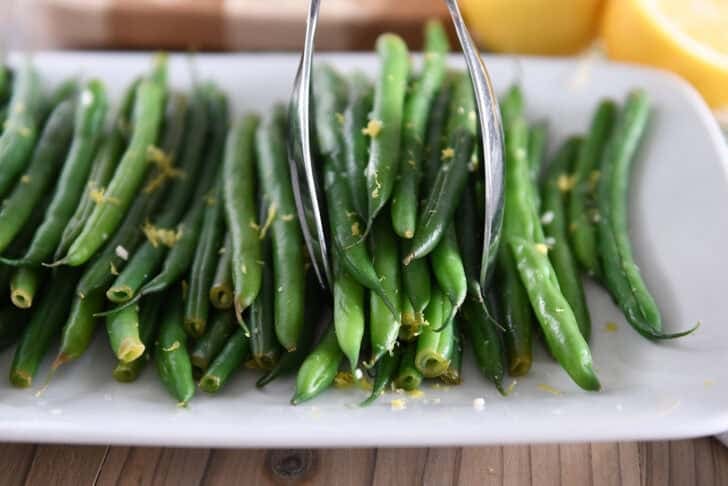 White tray with cooked green beans, lemon zest and tons scooping up portion of green beans.