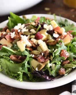 Green salad on white plate with roasted pumpkin seeds, chopped apples, candied pecans, feta cheese and red onions.
