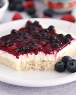 Square of white sheet cake topped with cream cheese layer and fruit topping on white plate with fresh blueberries.