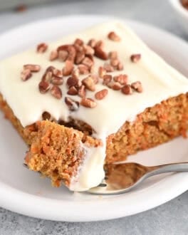 A piece of cream cheese frosted carrot sheet cake sprinkled with pecans on a white plate. A fork is taking a bite out of the cake.