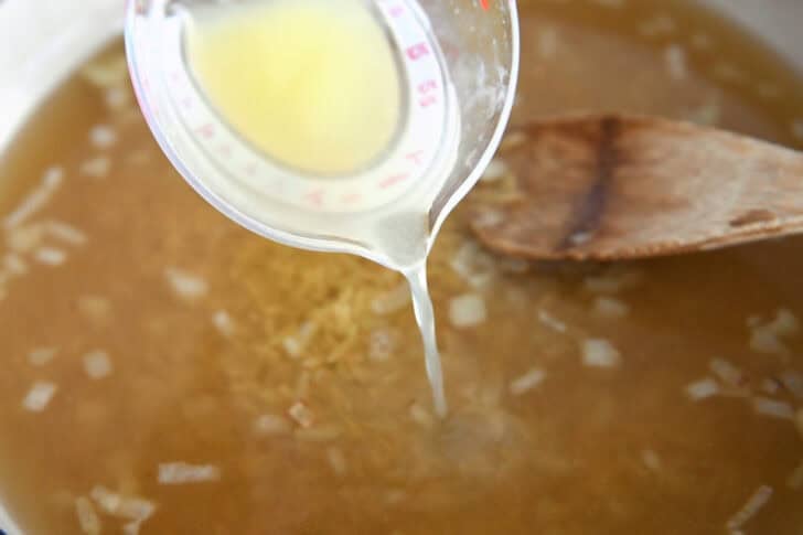 Pouring broth into white pot with onions and orzo.