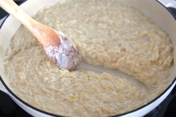 Wooden spoon scraping path through creamy orzo in white pan.