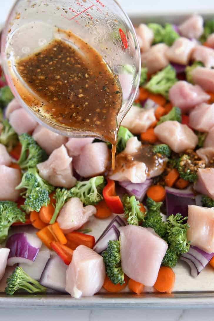 Pouring honey garlic sauce over chicken, broccoli, bell peppers, and carrots on sheet pan.