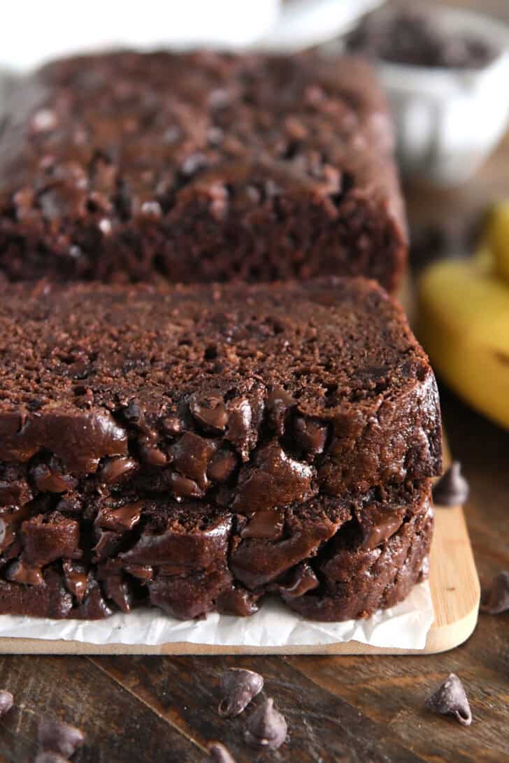 Two slices of chocolate banana bread stacked on parchment paper and wood cutting board.