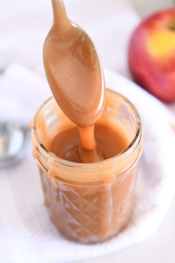 Spoon drizzling caramel sauce into glass jar with apple in the background.