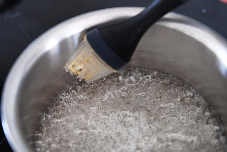Pastry brush brushing down sides of saucepan with simmering sugar and water.