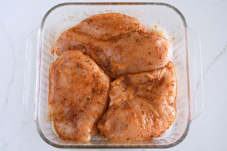 Three seasoned, uncooked chicken breasts in glass square pan.