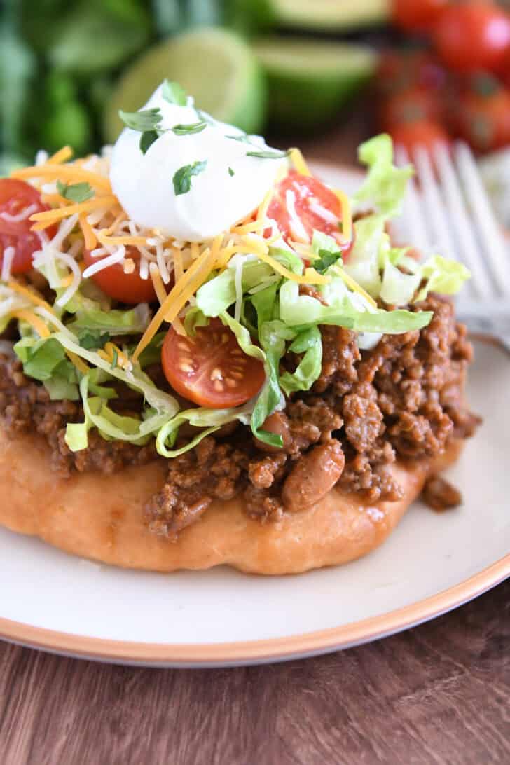 Piece of fry bread on cream plate topped with taco meat, lettuce, tomatoes, cheese, and sour cream.