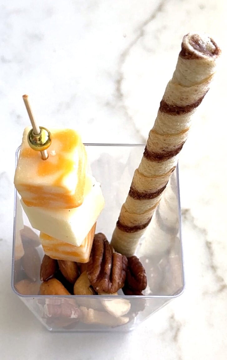 Square plastic cup with cracker, cheese skewer, and pirouline.