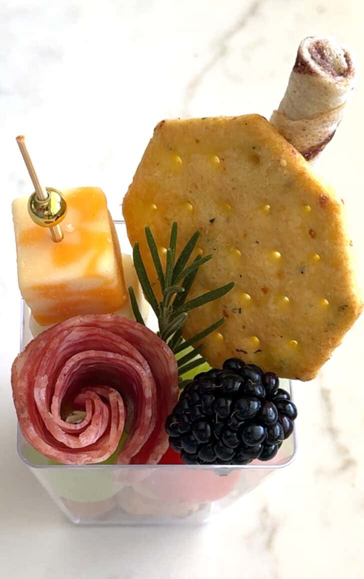 Square plastic cup with cracker, cheese skewer, rolled salami, blackberry, sprig of rosemary and pirouline.