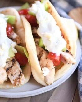 Two chicken gyros in flatbread with tzatziki, tomatoes and lettuce on gray plate.