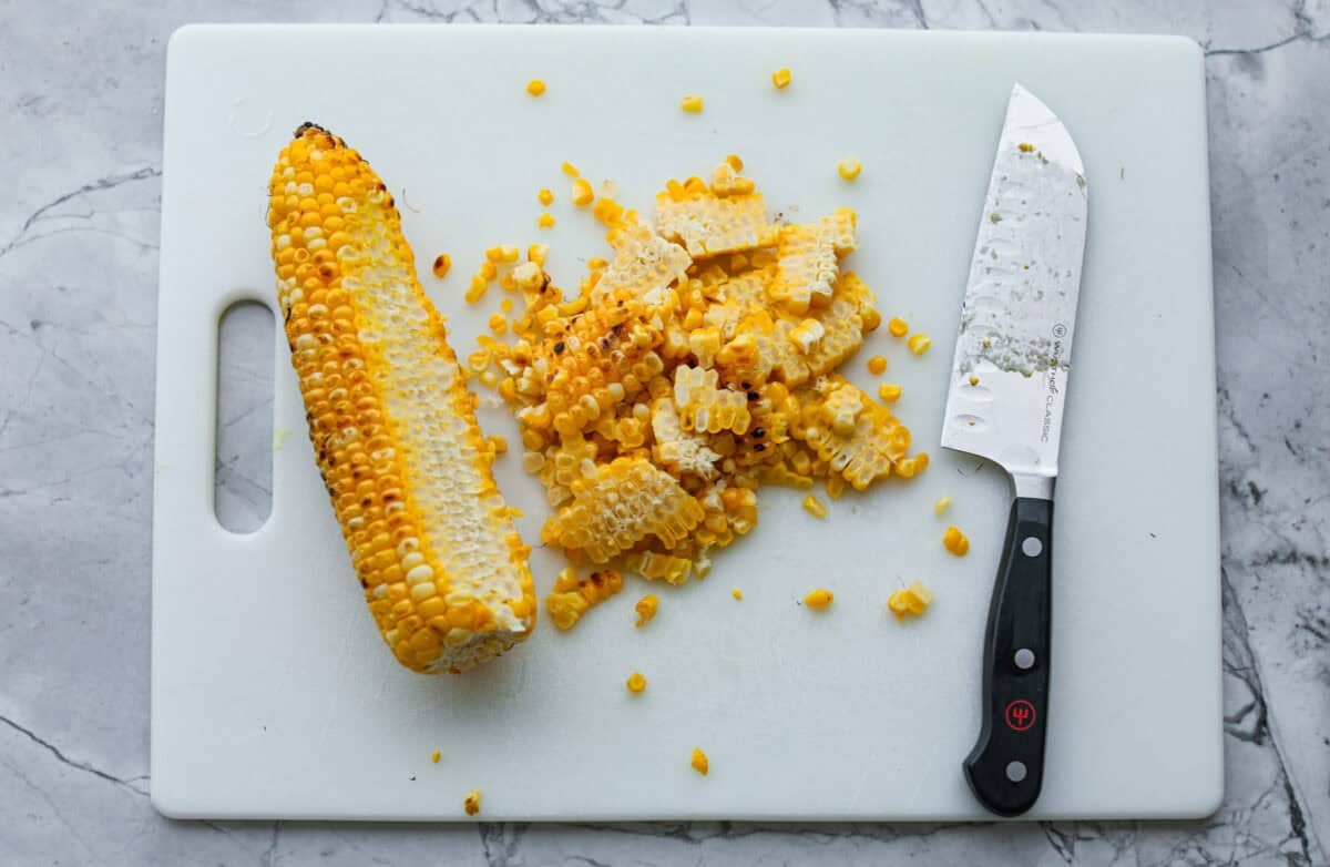Cutting board with a cob of corn with a pile of kernels cut off with a large knife. 