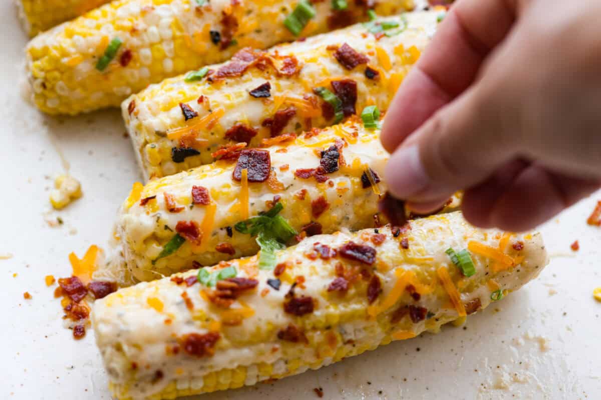 Someone sprinkling bacon, cheese, and green onions onto the corn. 