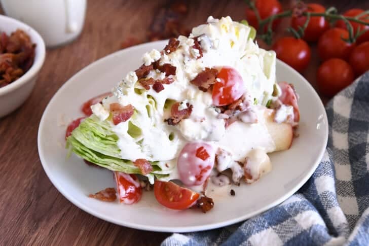 White plate with one iceberg wedge, blue cheese crumbles, blue cheese dressing, tomatoes, bacon, and sliced apples.