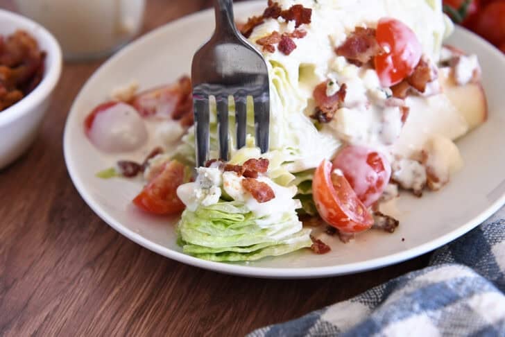 White plate with one iceberg wedge, blue cheese crumbles, blue cheese dressing, tomatoes, bacon, and sliced apples.